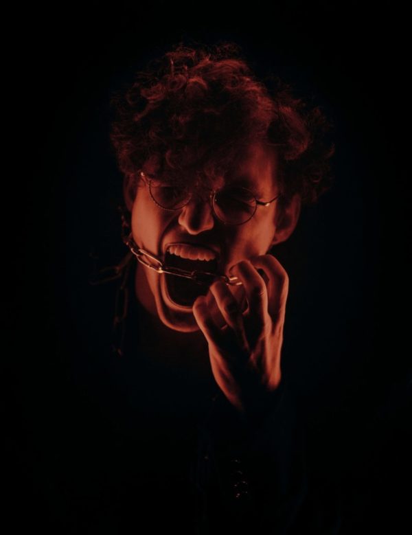 A white men cast in red light is yelling with a chain around his mouth